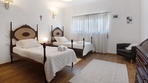 Une chambre à coucher avec deux lits en bois, une table de chevet, une commode, une lampe, une fenêtre avec des rideaux et un crucifix fixé au mur.