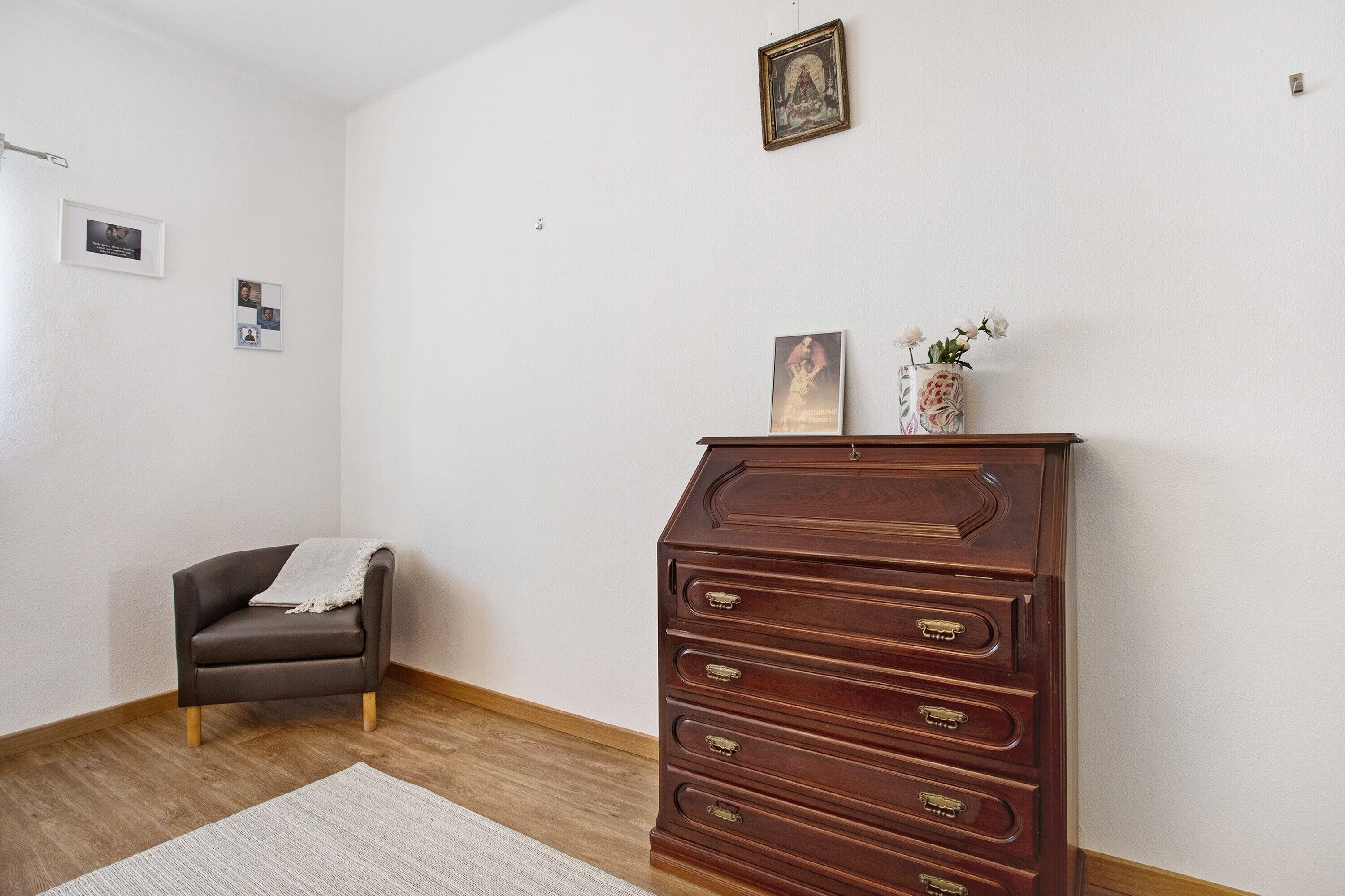 Une pièce comprenant une commode en bois, un fauteuil sombre, un petit tapis et des tableaux accrochés au mur.