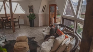 Une pièce chaleureuse avec un plafond haut, un plancher en bois, un canapé en osier agrémenté de coussins, une table avec des chaises, une plante en pot et de grandes fenêtres offrant une vue sur l’extérieur.