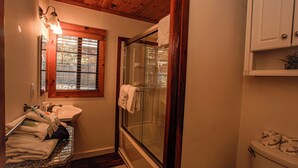 A bathroom with a glass-enclosed shower, a sink with a countertop, and a window with blinds. - Fox Cabin (Big Bear City)