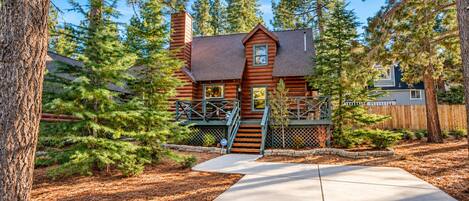 A log cabin with a wooden deck, a chimney, and a fenced yard surrounded by pine trees.