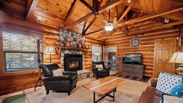 A log cabin interior with a stone fireplace, leather furniture, a television, and a ceiling fan. - Fox Cabin (Big Bear City)