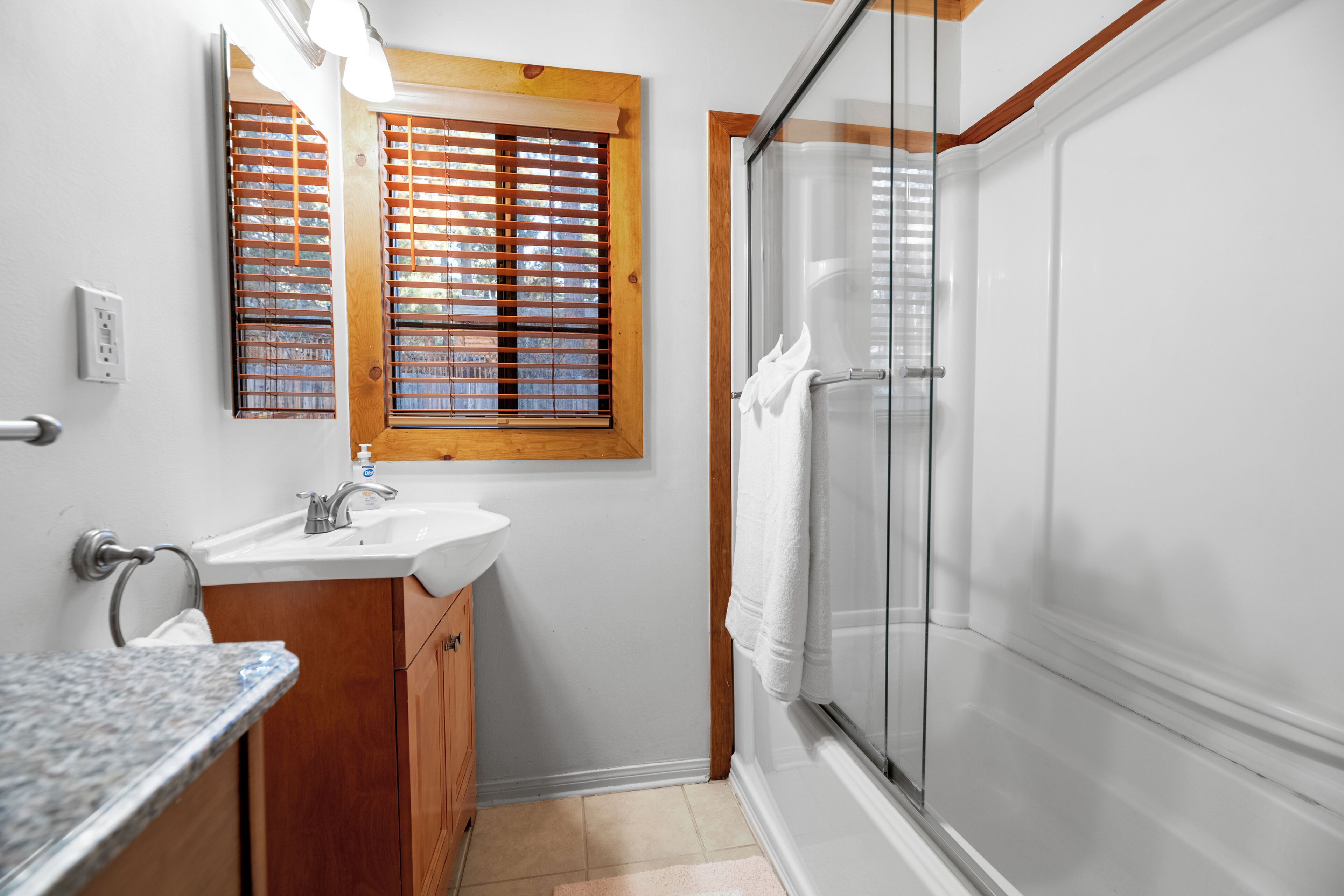 Ein Badezimmer mit Waschbecken, Spiegel, Fenster mit Jalousien und einer Duschkabine aus Glas.