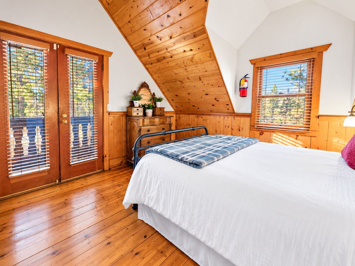 A bedroom with a wooden ceiling, a bed with a white and blue bedspread, a dresser, and a window with blinds.