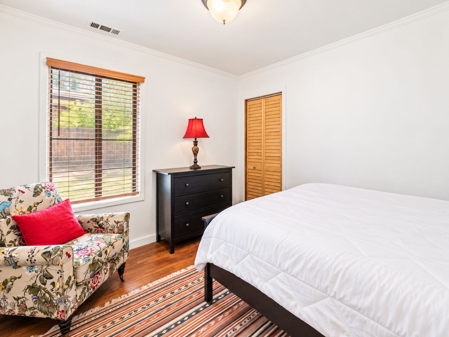 A bedroom with a bed, a dresser, a chair, and a window with blinds.