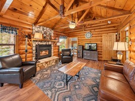 A log cabin interior with a stone fireplace, leather furniture, and a television.