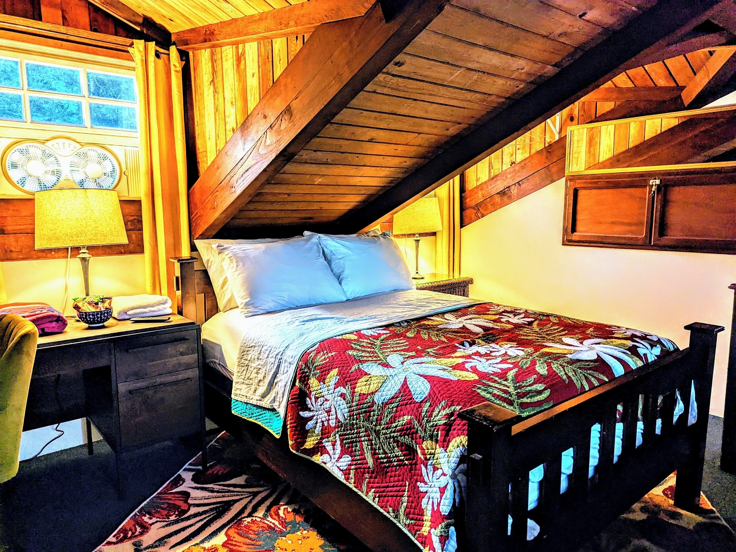 A cozy attic bedroom with a wooden ceiling, a bed with a floral quilt, a nightstand with a lamp, and a window with curtains.