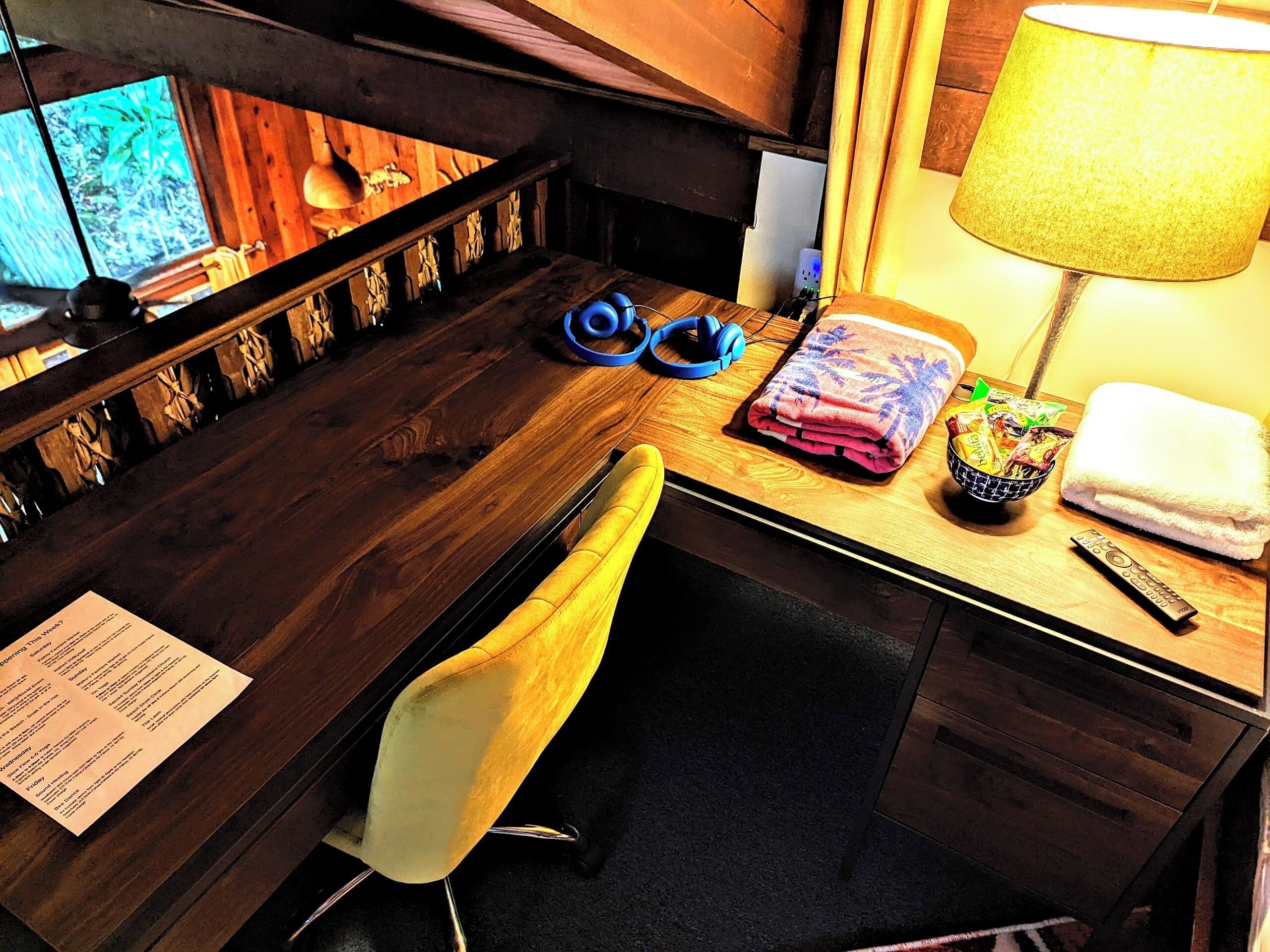 Une chambre d’hôtel avec un bureau en bois, une chaise, une lampe, un casque audio, une serviette, une télécommande et un menu.