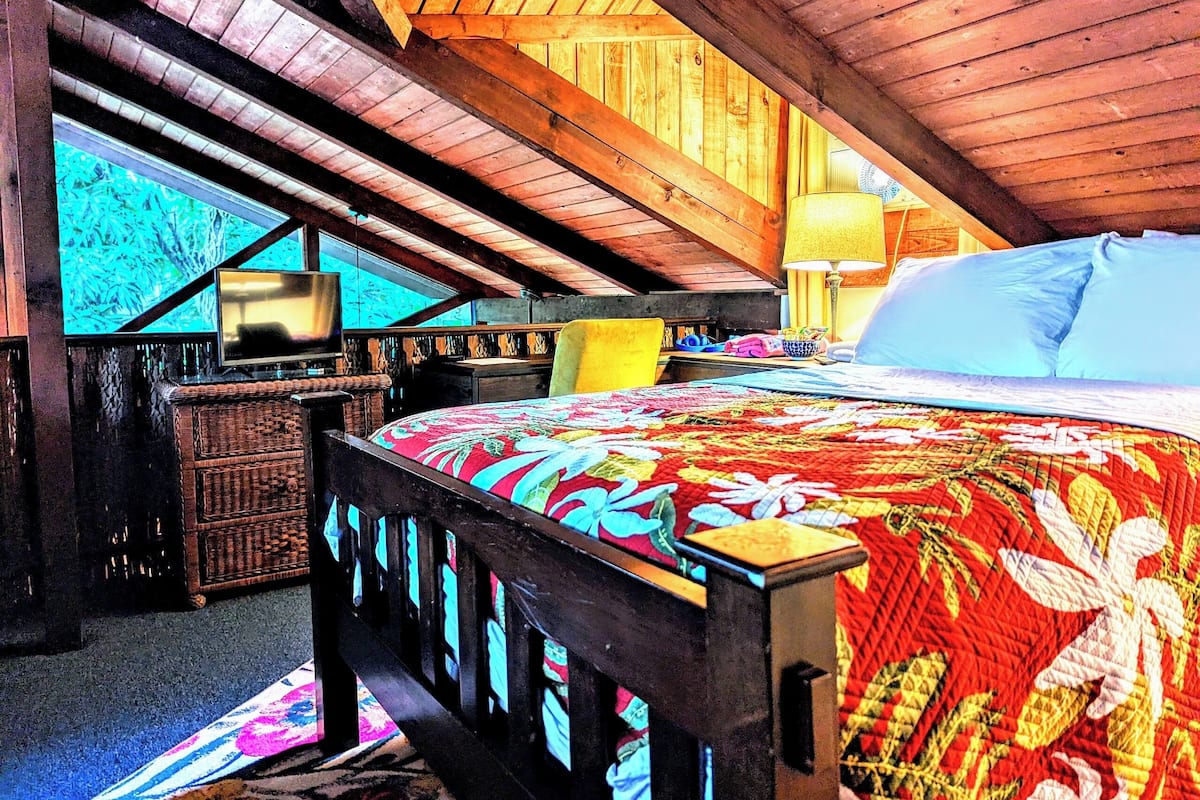 A cozy attic bedroom with a wooden ceiling, a bed with a colorful quilt, a desk with a lamp, and a television.