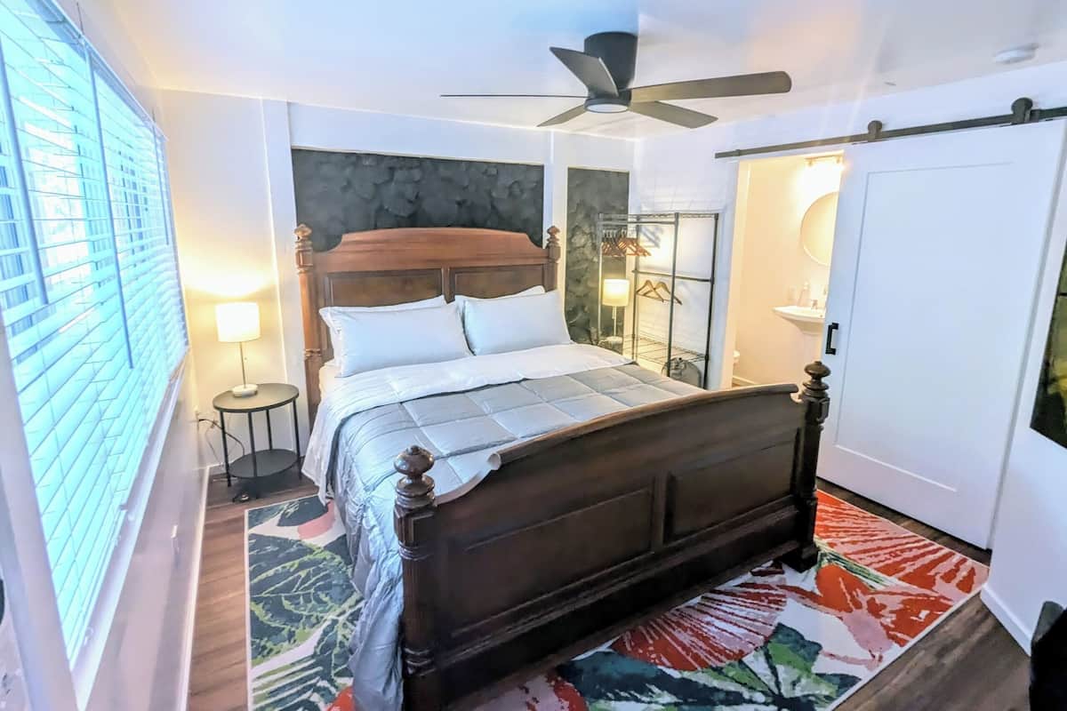 A bedroom with a wooden bed, a ceiling fan, a stone accent wall, a small bedside table with a lamp, and a colorful rug.