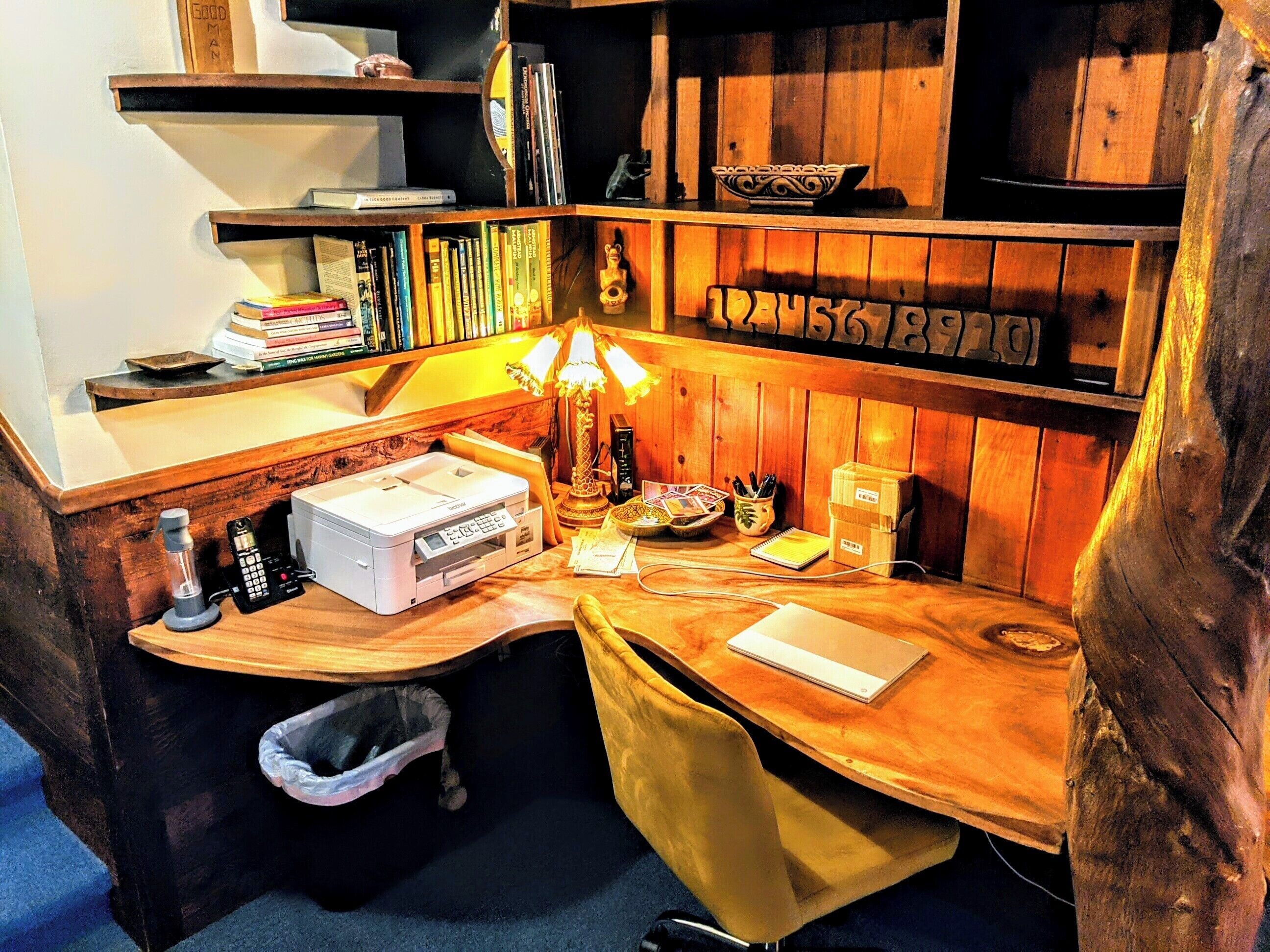 Un espace de travail accueillant, avec un bureau en bois, une lampe, une imprimante et une bibliothèque remplie de livres et d’objets décoratifs.