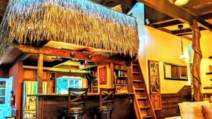 A rustic interior with a thatched roof, wooden bar, stools, and a staircase.