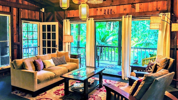 A cozy living room with a sofa, coffee table, and armchair, wooden walls, and large windows with curtains.