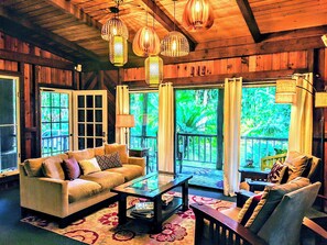 A cozy living room with a sofa, coffee table, and armchair, wooden walls, and large windows with curtains. - Kehena Mauka Nui LGBTQ Clothing Optional (Pahoa)