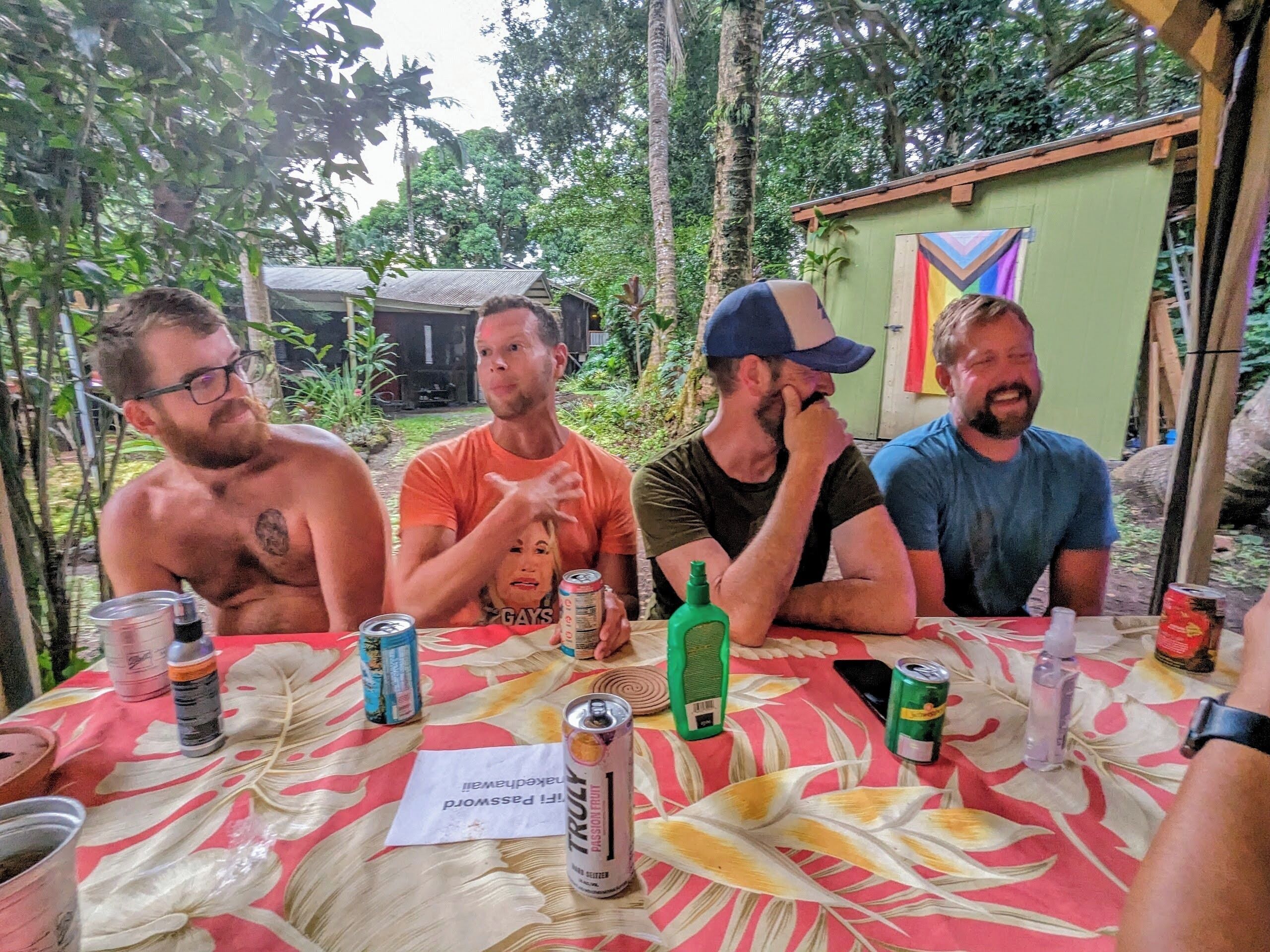 Four people seated at an outdoor table with a floral tablecloth, surrounded by various beverages and a small green bottle.