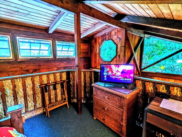 A rustic cabin interior with wooden walls, a television displaying 'Big Mouth', and a large window with a view of greenery.