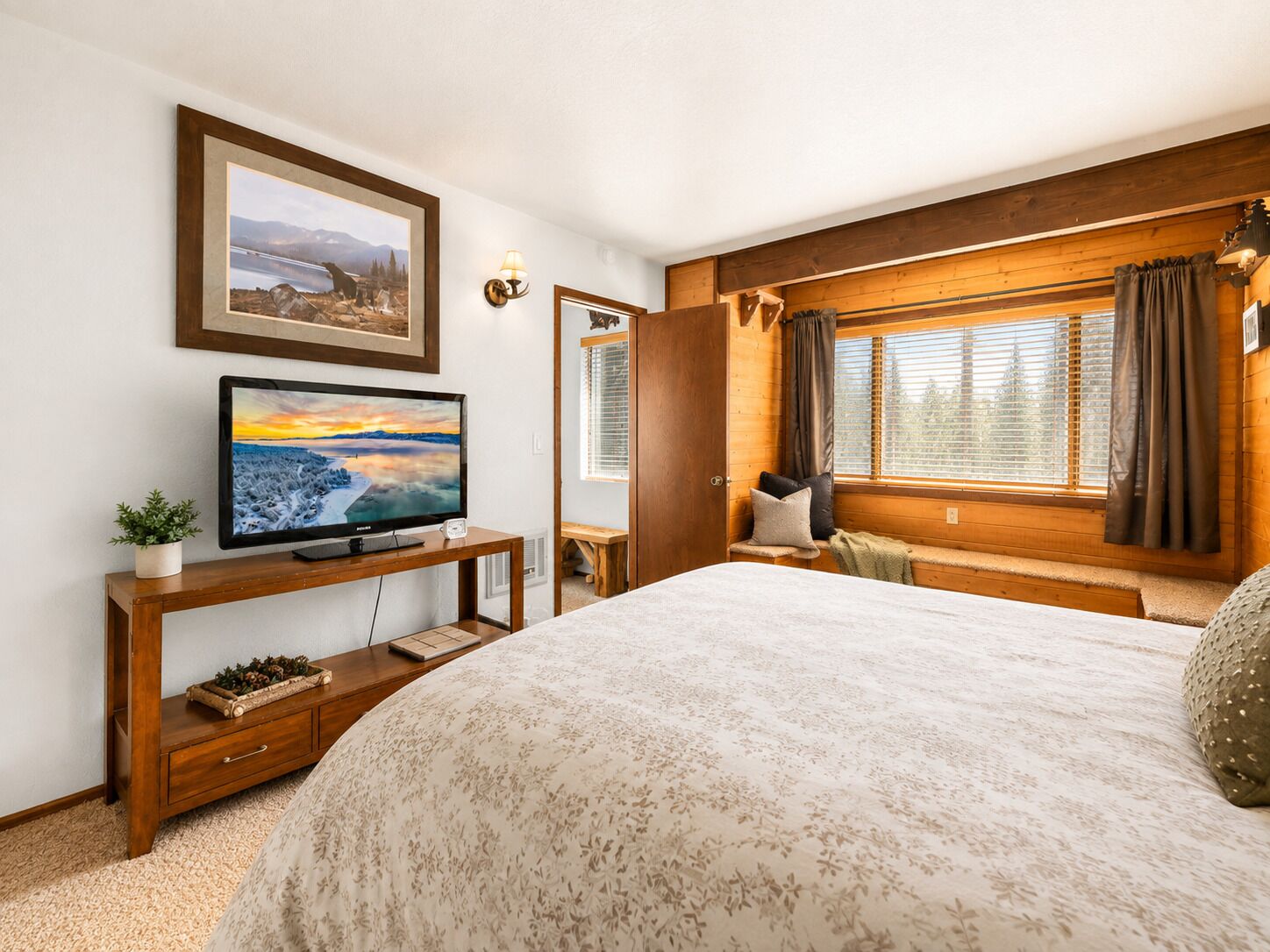 A bedroom with a bed, a television, a window with curtains, a wooden headboard, and a picture on the wall.