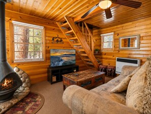 A cozy cabin interior with a fireplace, wooden walls, a sofa, a television, and a staircase. - Highland Hideaway (Big Bear Lake)