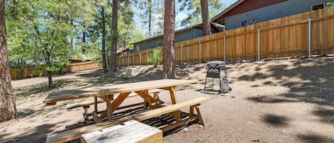 A picnic area with wooden benches and a grill, surrounded by trees and a wooden fence.