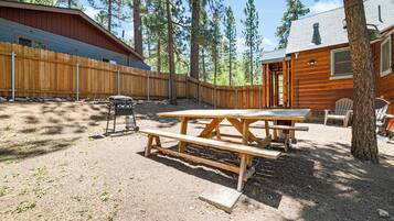 Una cabaña rústica con terraza de madera, mesa de picnic y zona de estar al aire libre, rodeada de altos pinos.