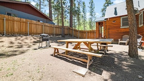 Een rustieke hut met een houten terras, picknicktafel en zitgedeelte buiten, omringd door hoge dennenbomen.