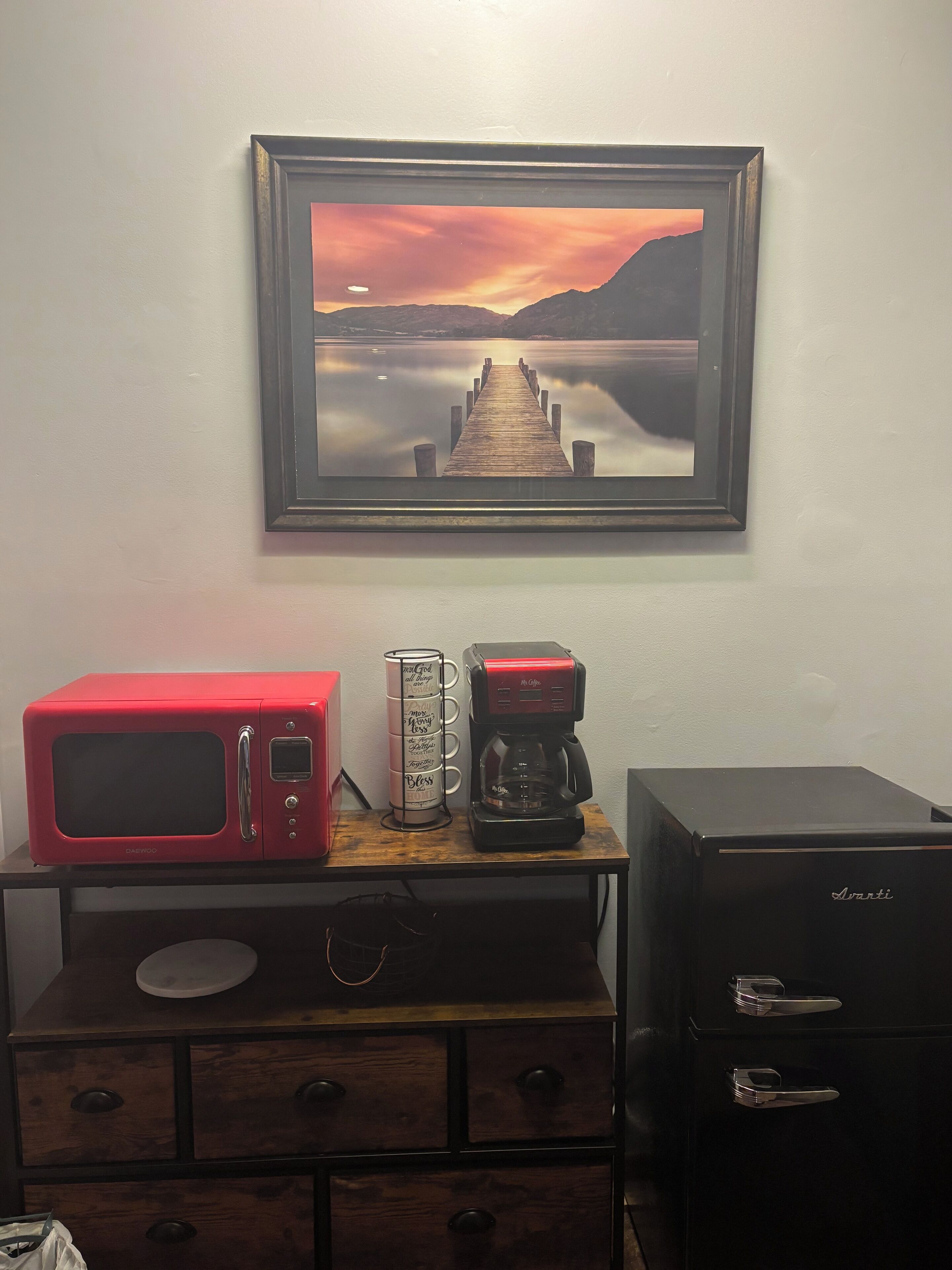 Een kitchenette met een magnetron, koffiezetapparaat en een lijst met een zonsondergang boven een meer.