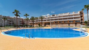 Une piscine entourée d’une terrasse en briques, bordée de palmiers et d’un immeuble à plusieurs étages doté de balcons.