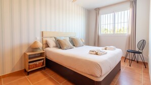 Une chambre à coucher comprenant un lit, une table de chevet, une chaise, une fenêtre avec des rideaux et un ventilateur de plafond.