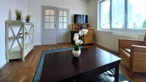 A living room with a dark wooden coffee table, a white console table with plants, a wicker chair, a television on a wooden stand, and a large window with a view of the outside.