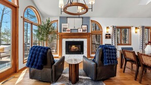 A cozy living room with a fireplace, leather chairs, a dining table, and large windows. - Hot Tub | Sleeps 20 | Private Beach | Game Room (Two Rivers)