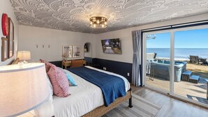 A bedroom with a large bed, a ceiling with a wave pattern, a chandelier, a view of the beach through a sliding door, and a hot tub outside. - Hot Tub | Sleeps 20 | Private Beach | Game Room (Two Rivers)