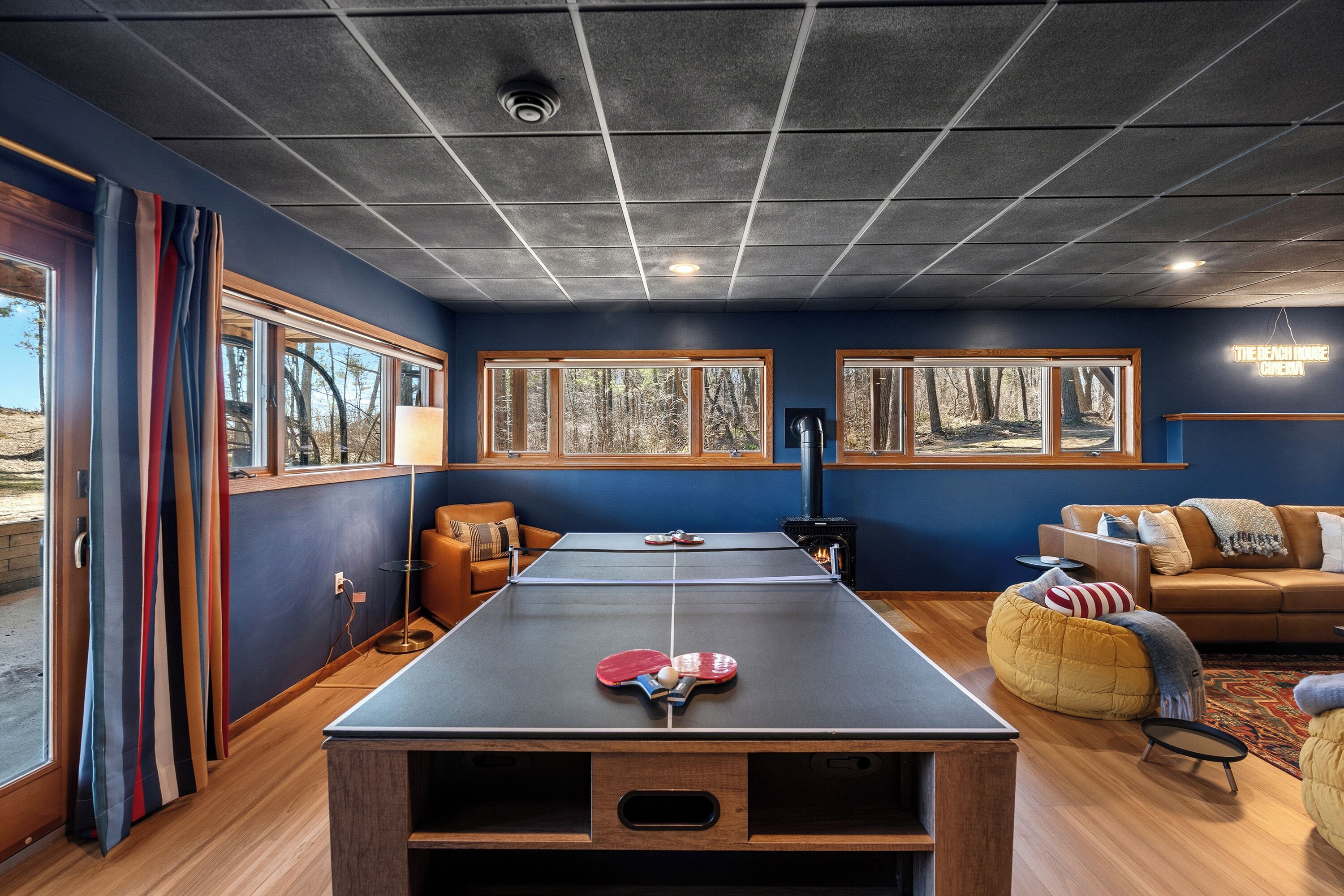 A room with a ping pong table, two couches, a fireplace, and large windows with forest views.