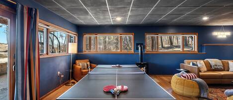 A room with a ping pong table, two couches, a fireplace, and large windows with forest views.