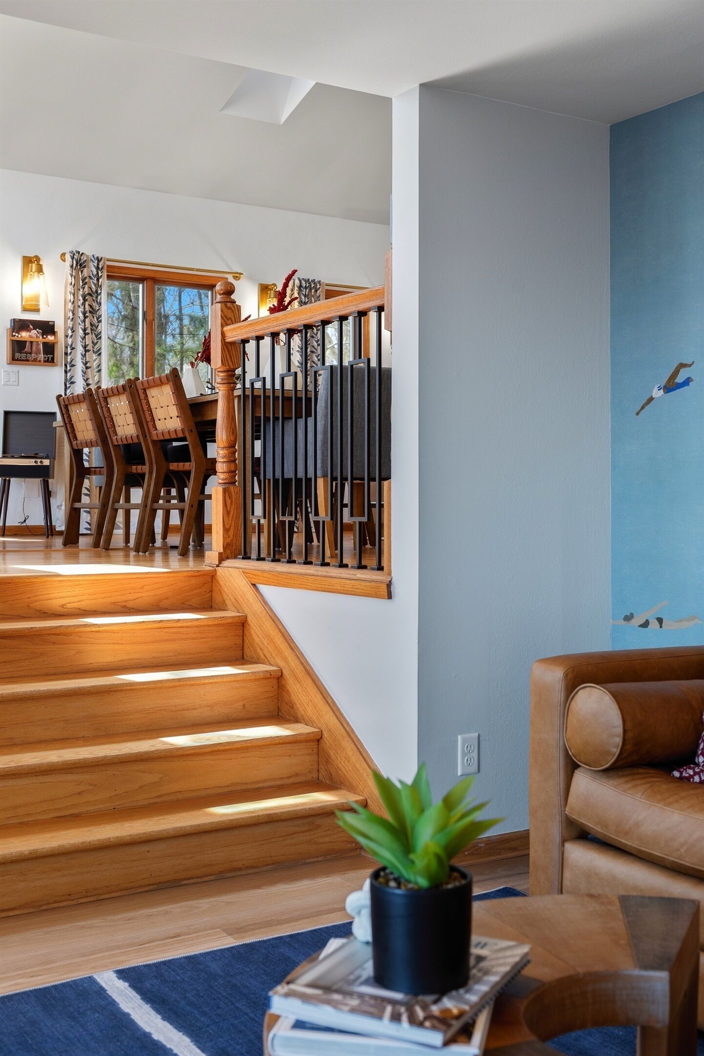 Un escalier en bois mène à un coin repas avec des chaises et une table, un mur bleu orné d’une fresque représentant une mouette, et un canapé en cuir.