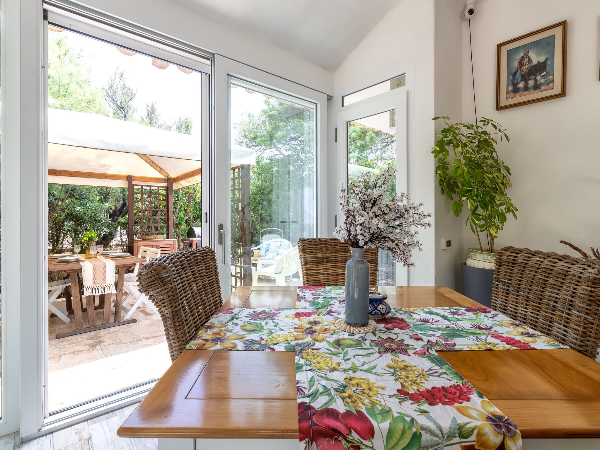 A dining area with a wooden table, wicker chairs, and a floral tablecloth. There is a potted plant on the table and a view of the outdoors through glass doors.
