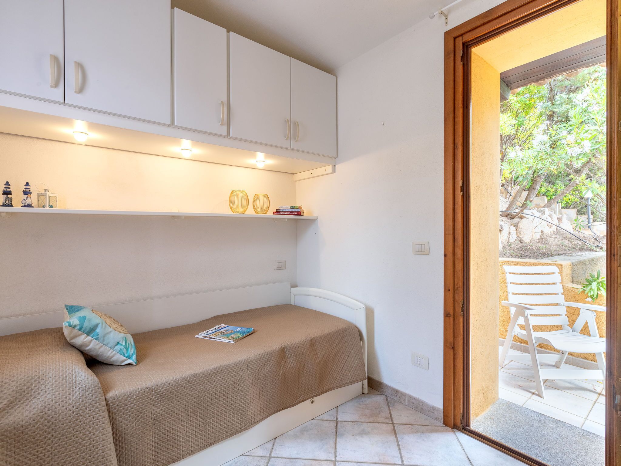 A small, cozy room with a single bed, a shelf with decorative items, and a door leading to an outdoor seating area.