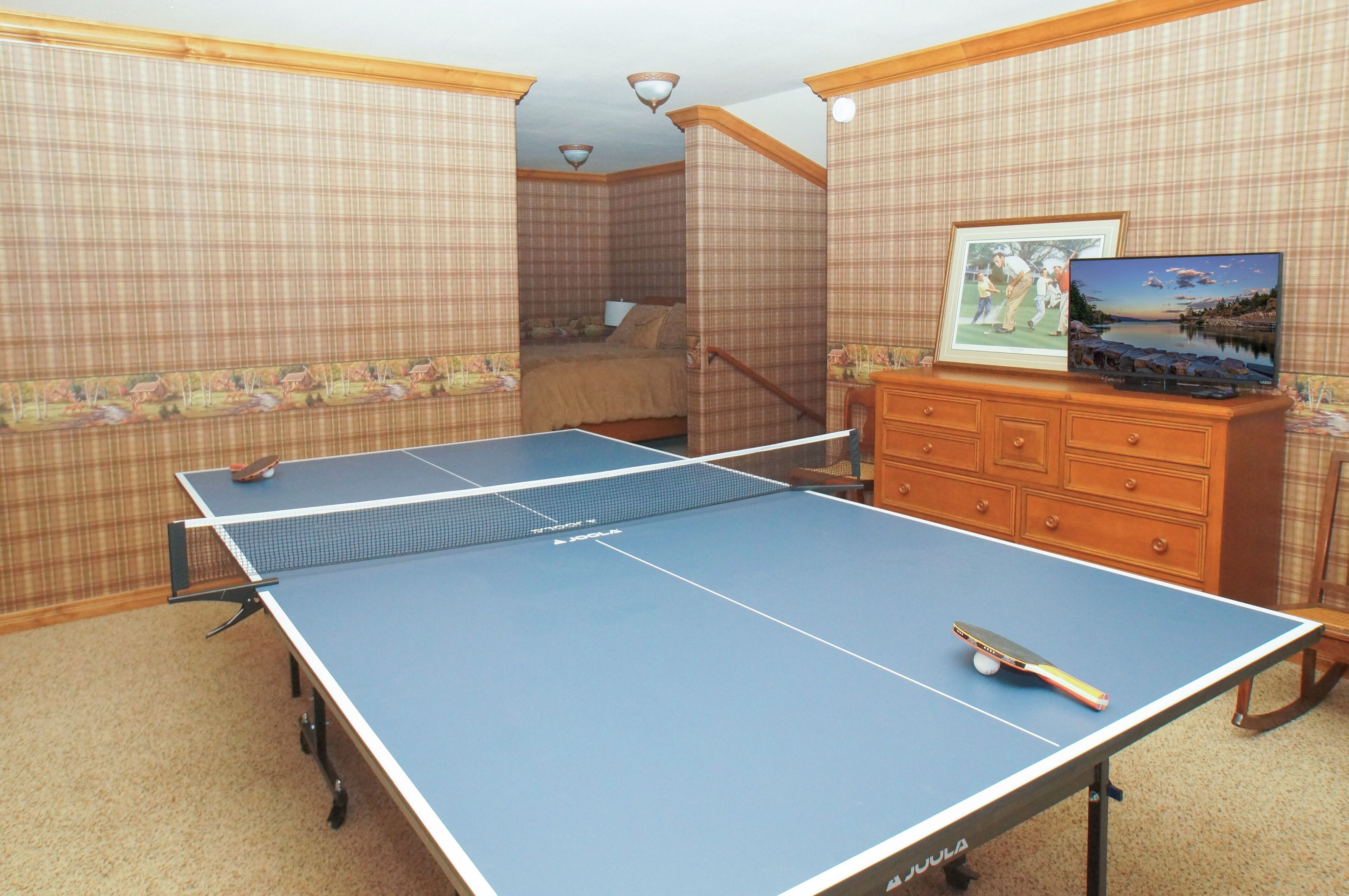 A room with a ping pong table, a dresser, a framed picture, and a bed with a headboard.