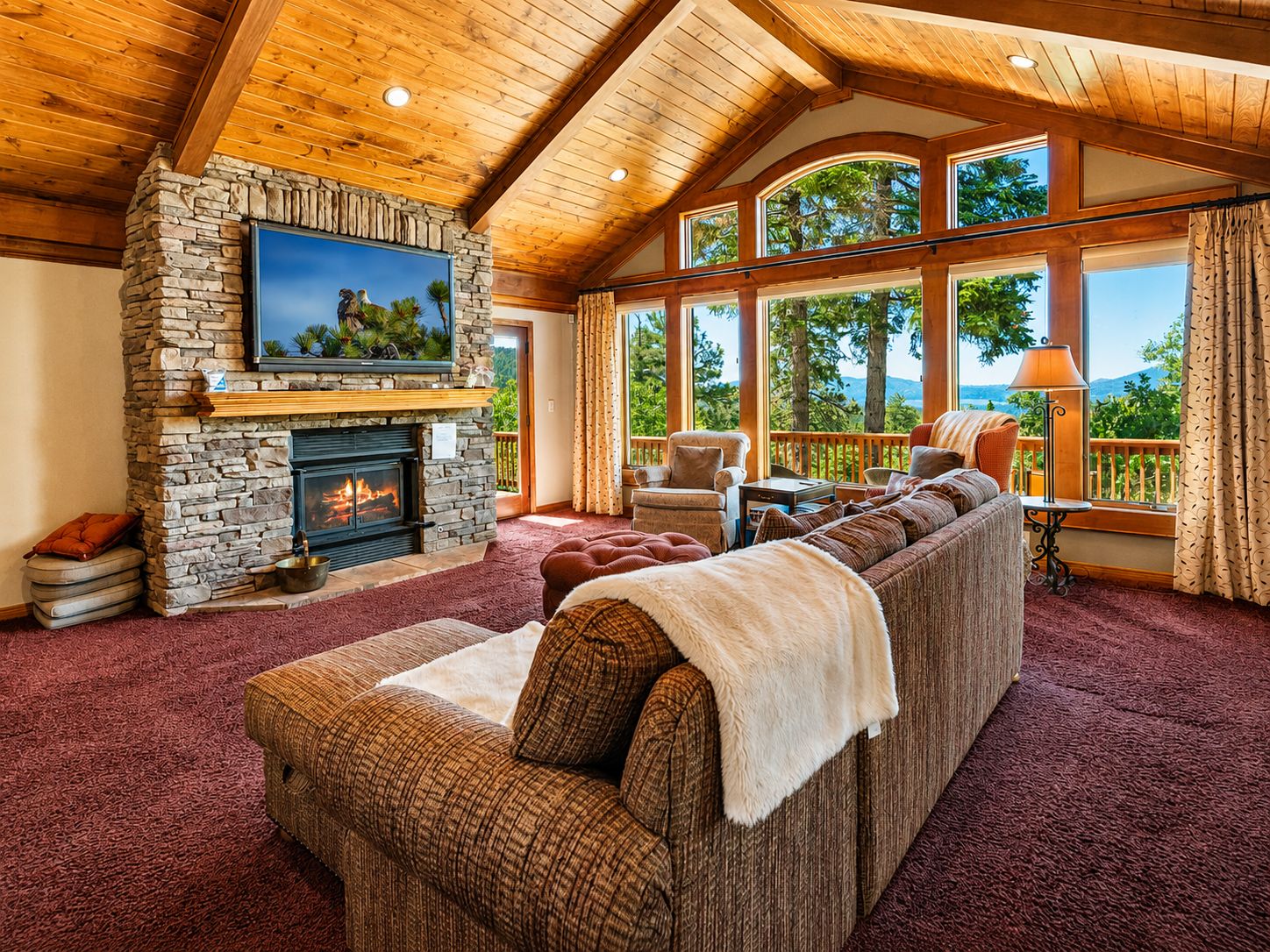 A cozy living room with a stone fireplace, a large flat-screen TV, a sectional sofa, and a view of the outdoors through large windows.