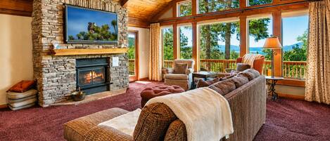 A cozy living room with a stone fireplace, a large flat-screen TV, a sectional sofa, and a view of the outdoors through large windows.