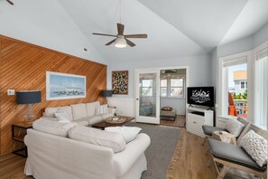 A modern living room with a sectional sofa, a ceiling fan, a television, and a view of the outdoors. - Blue Pearl: Steps to the Beach, Ocean Views, Spacious Decks (Nags Head)