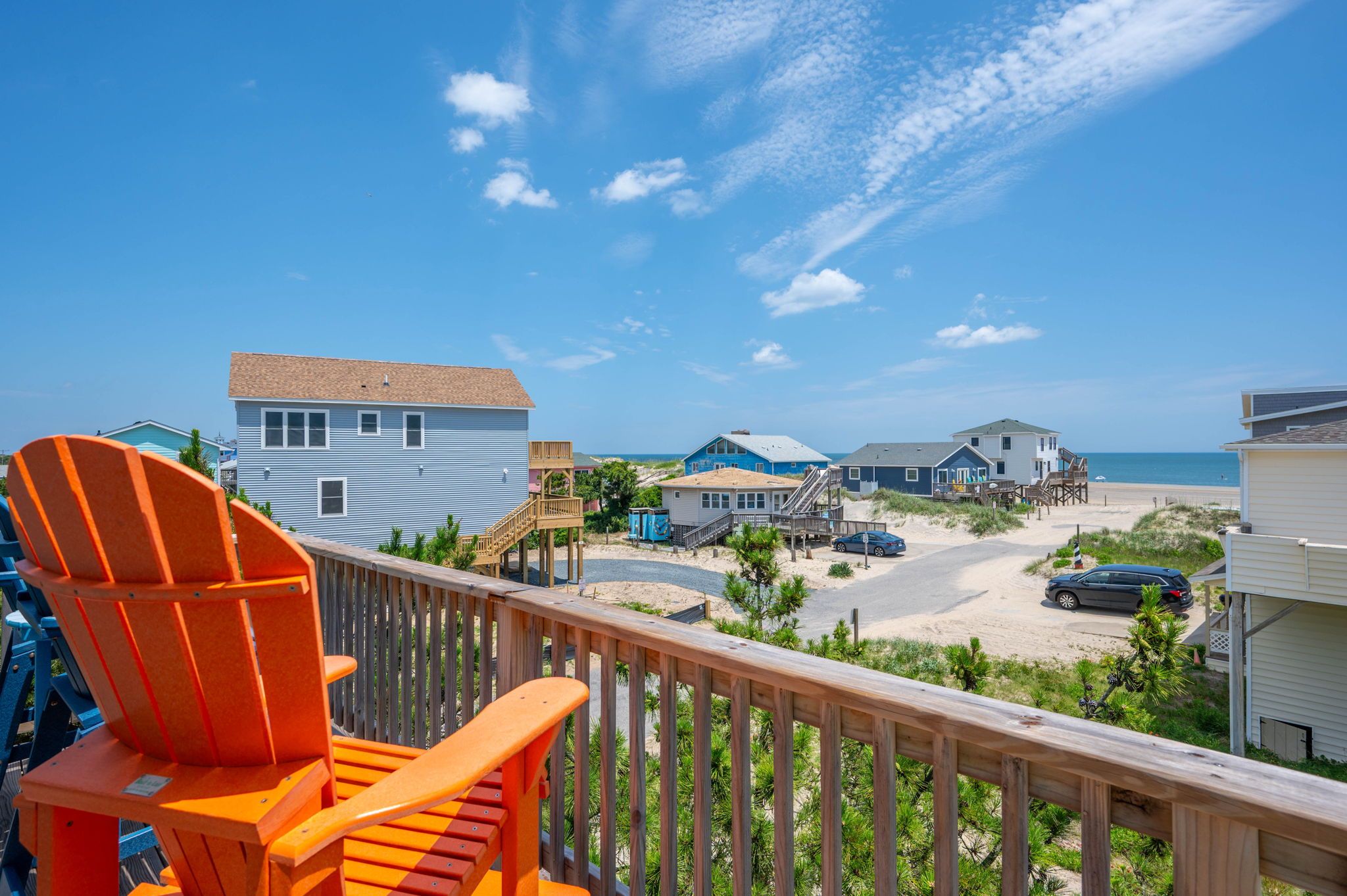 Vue sur la plage depuis un balcon doté d’une terrasse en bois, avec une chaise, un bâtiment au toit bleu et une plage de sable.