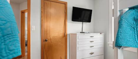 A room with a wooden door, a white dresser with drawers, a flat-screen TV mounted above it, and a bunk bed with a blue blanket.