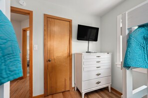 Une pièce avec une porte en bois, une commode blanche avec des tiroirs, un téléviseur à écran plat fixé au-dessus, et un lit superposé avec une couverture bleue.
