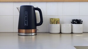 Een moderne keuken met een zwarte waterkoker, met de aanduiding 'Russell Hobbs', en drie witte bakjes met de tekst 'Koffie', 'Thee' en 'Suiker'.