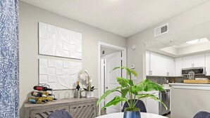 Une cuisine moderne avec des meubles blancs, un four à micro-ondes et un évier. On y trouve un coin repas avec une table ronde et des chaises, ainsi qu’une plante en pot posée sur la table.