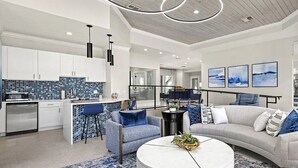 Une cuisine moderne avec des meubles blancs et un dosseret à motifs bleus, un coin repas avec un canapé gris et des coussins bleus, et un salon avec un canapé gris et une table basse blanche.