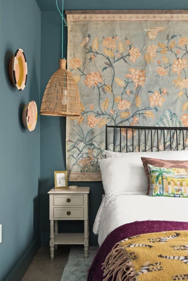 Un dormitorio con una cama, una mesita de noche, una lámpara de mimbre y un tapiz floral en la pared.
