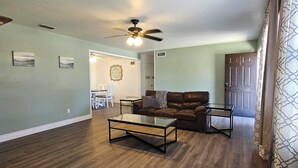 A spacious living room with hardwood flooring, a glass coffee table, a brown leather sofa, and a ceiling fan.