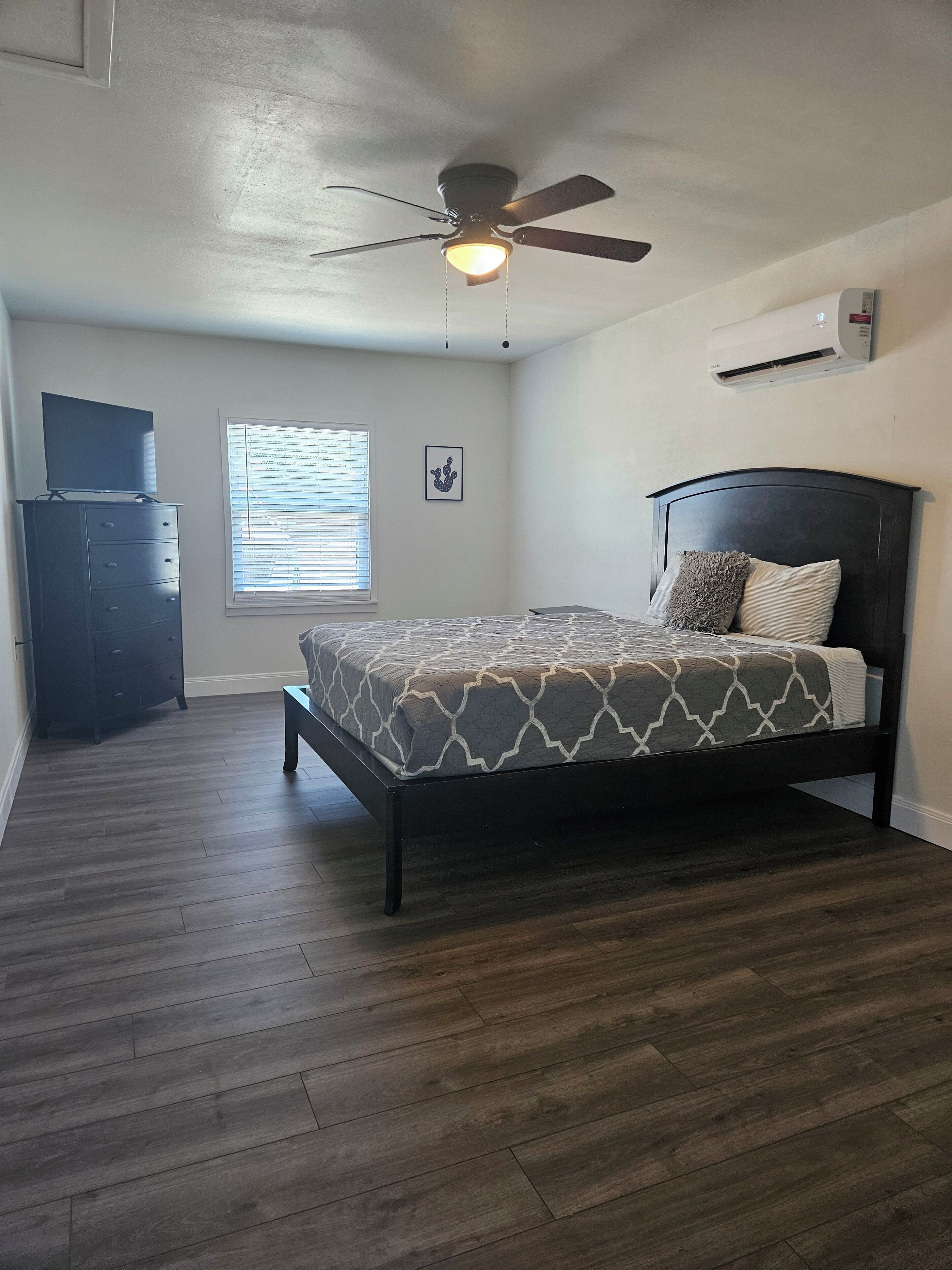 Une chambre à coucher comprenant un lit, un ventilateur de plafond, une commode, une fenêtre avec des stores et un appareil de climatisation.