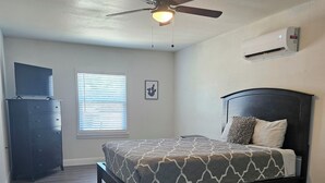 A bedroom with a bed, a ceiling fan, a dresser, a window with blinds, and an air conditioning unit.